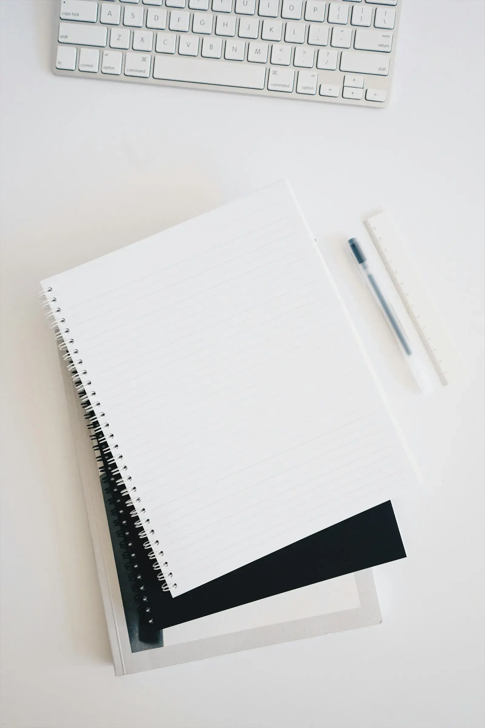 A blank spiral notebook and a pen on a white desk beside a keyboard.