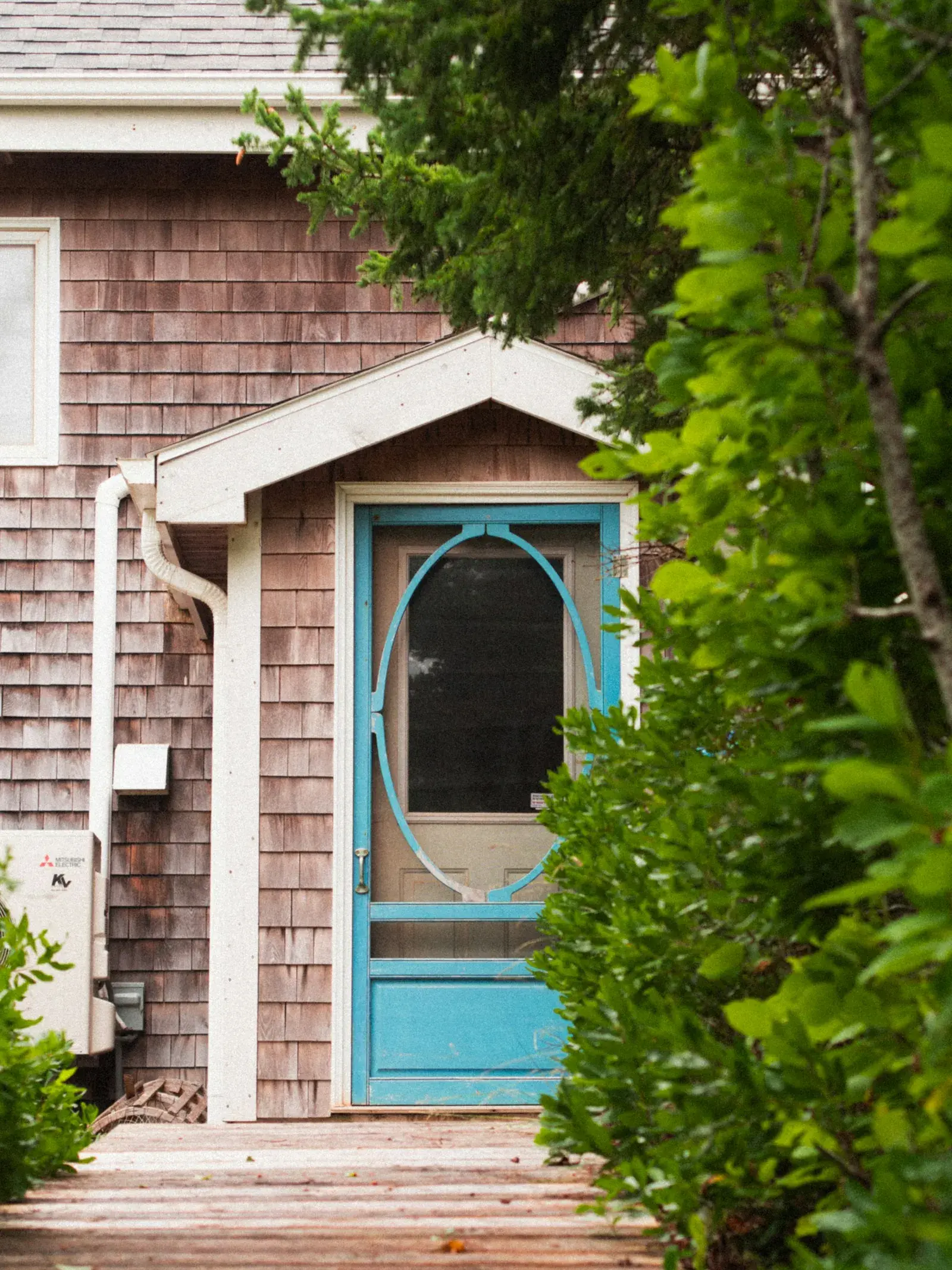 A cottage with brown wooden shingles and a blue screen door, with green leaves in front.