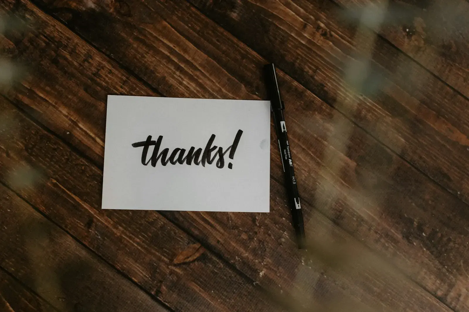 A white card with the word thanks written on it, lying next to a black pen on a dark wooden table.
