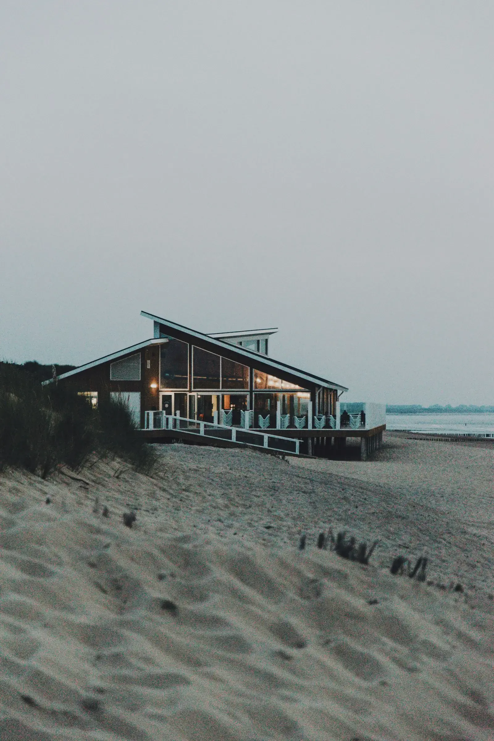 A modern holiday house on a sandy beach at dusk, with warm light in the windows.